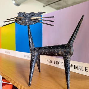 May include: A black metal cat sculpture with long whiskers and a tall, pointed tail. The sculpture is on a wooden shelf with the word "TURBO" and "PERIFUCK VINKLE" visible in the background.