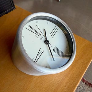 Puede incluir: Un reloj de escritorio plateado y redondeado con esfera blanca y números romanos negros. El reloj tiene una cubierta de vidrio y está sobre una superficie de madera. La esfera del reloj tiene las letras "Z&R" impresas.