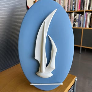 May include: A light blue oval wall art piece featuring a white abstract design. The artwork is displayed on a wooden surface and has a white support bar at the bottom. The background shows a room with books.