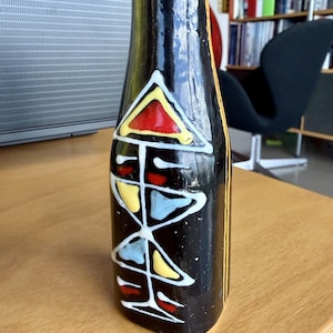 May include: A black ceramic bottle with abstract geometric designs in red, yellow, blue, and white. The bottle has a narrow neck and a rounded base. Vertical yellow stripes accent the sides.