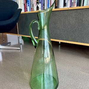 Pode incluir: Uma jarra alta de vidro verde com uma alça e um bico largo. A jarra tem formato cônico e um detalhe dourado perto do topo. Está sobre um chão claro. O fundo inclui uma estante cheia de livros.