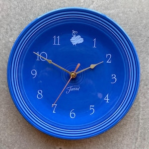 Puede incluir: Un reloj de pared redondo azul cobalto con números blancos y manecillas doradas. La esfera del reloj presenta la palabra "Fiesta" y un emblema decorativo. El reloj tiene un borde acanalado.