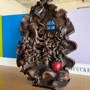 May include: A detailed, brown sculpture of a whimsical treehouse with a blue window and a red apple. The artwork features intricate details of leaves and branches, creating a fairytale aesthetic. The sculpture is set on a black metal base.