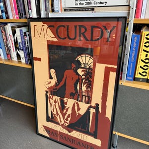 May include: A framed art print with the name "MCCURDY" in bold letters. The artwork uses red, black, and beige tones, showing a figure in a room with a bed and a window. The text "OBRAS SANJUANERAS" is at the bottom of the print.