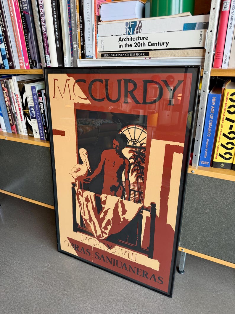 May include: A framed art print with the name "MCCURDY" in bold letters. The artwork uses red, black, and beige tones, showing a figure in a room with a bed and a window. The text "OBRAS SANJUANERAS" is at the bottom of the print.