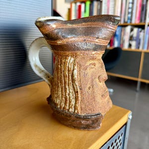 Puede incluir: Una taza de cerámica artesanal con un diseño de rostro único. La taza presenta una combinación de colores marrón y beige con un asa blanca. El diseño de la taza incluye una superficie texturizada y una forma artística distintiva.