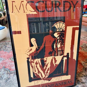 May include: Framed art print with the name "MCCURDY" in tan. The artwork shows a man, a pelican, and a window scene with the text "OBRAS SANJUANERAS" and Roman numerals. The color scheme includes tan, red, and black, creating a striking visual.