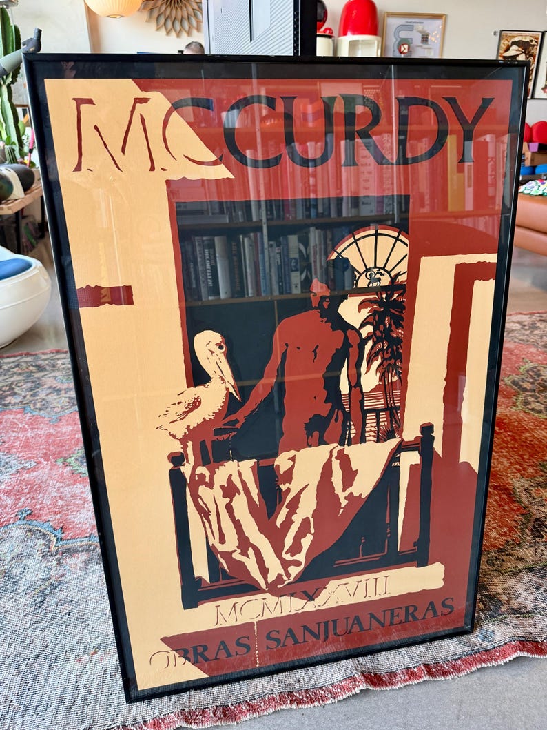 May include: Framed art print with the name "MCCURDY" in tan. The artwork shows a man, a pelican, and a window scene with the text "OBRAS SANJUANERAS" and Roman numerals. The color scheme includes tan, red, and black, creating a striking visual.