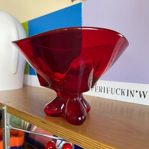 Może przedstawiać: Żywy, czerwony szklany wazon o unikalnym, rzeźbiarskim designie. Wazon ma rozszerzoną krawędź i spoczywa na trzech zaokrąglonych, łzowatych nóżkach. Przezroczyste szkło przepuszcza światło i jest wystawione na drewnianej półce.