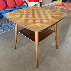 May include: A vintage wooden chess table with a checkerboard top. The table has a lower shelf and four tapered legs with brass-colored tips. The tabletop is approximately 61 cm square.
