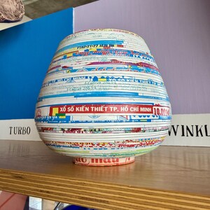 May include: A decorative, bulbous vase made from layered paper strips. The strips are multi-colored with text and patterns, including the words "HO CHI MINH" and "TURBO". The vase sits on a wooden shelf.