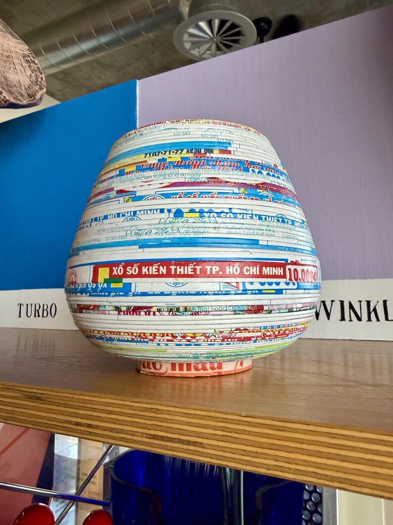 May include: A decorative, bulbous vase made from layered paper strips. The strips are multi-colored with text and patterns, including the words "HO CHI MINH" and "TURBO". The vase sits on a wooden shelf.