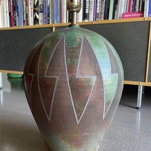 May include: A decorative table lamp with a bulbous, vase-like base. The base features a gradient of colors, including brown, green, and copper, with silver lightning bolt designs. The lamp has a brass-colored neck and a standard lamp socket.