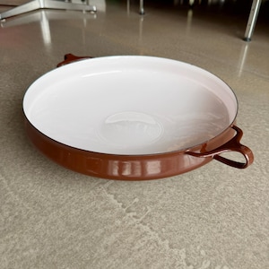 May include: A round, shallow baking dish with a glossy brown exterior and a smooth white interior. The dish features two small, curved loop handles on opposite sides, constructed from a solid material with a reflective, enameled finish.