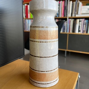 Puede incluir: Un jarrón de cerámica blanco con rayas marrones y naranjas. El jarrón tiene una textura moteada y una parte superior ligeramente acampanada.