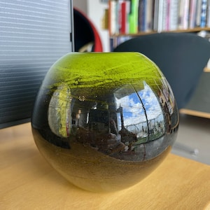 Può includere: Un vaso rotondo in vetro con una sfumatura di colori. La parte superiore è verde brillante, che sfuma in nero nella parte inferiore. Il vaso riflette l'ambiente circostante, tra cui un cielo blu e nuvole.