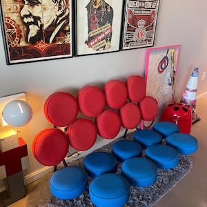 May include: A contemporary interior with a red and blue seating arrangement. The seating features round, red-upholstered backrests and blue, circular seats. The furniture is placed on a grey rug, with framed artwork displayed on the wall behind.