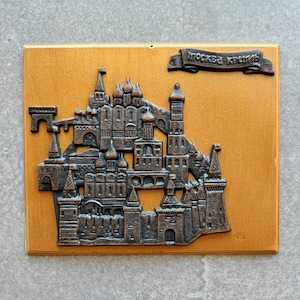 May include: A rectangular wooden wall plaque featuring a bronze-colored metal relief of the Moscow Kremlin. The relief depicts towers, walls, and onion domes. A small banner at the top right corner contains the Cyrillic text "МОСКВА-КРЕМЛЬ".