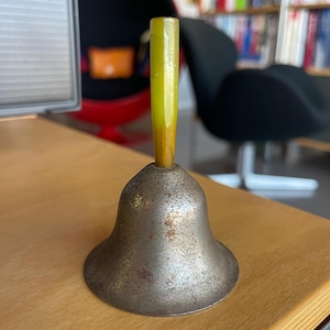 May include: A small, silver bell with a worn, aged finish. The bell has a yellow plastic handle.