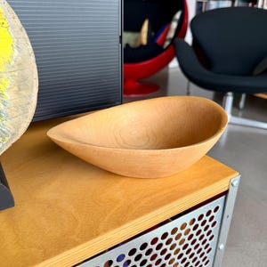 May include: A light-colored, smooth wooden bowl with an asymmetrical, teardrop-shaped rim. The bowl features a natural wood grain pattern visible on its interior surface and a curved, shallow basin design, resting on a flat surface.
