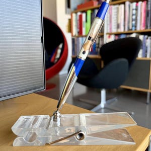 May include: A pen with a blue and silver barrel is displayed in a clear plastic clothespin-shaped pen holder. The pen holder sits on a wooden desk. The pen has a clear barrel with a design inside.