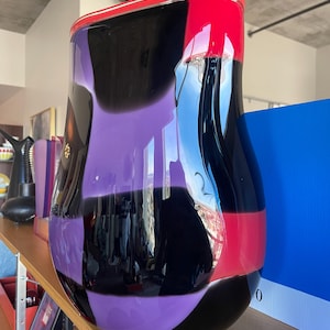 Könnte beinhalten: Eine große, farbenfrohe Glasvase mit einem geometrischen Muster in Schwarz, Lila und Rot. Die Vase ist glänzend und reflektiert das Licht.