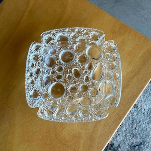 Peut inclure: Un cendrier carré en verre transparent présentant une surface texturée avec des motifs circulaires en relief semblables à des bulles de tailles variées. Le verre a une apparence épaisse et lourde avec un design en relief créant une finition bosselée.