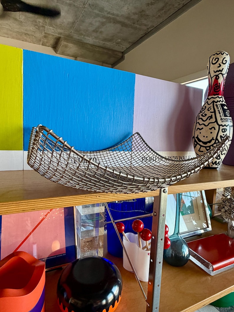 May include: A curved, rectangular wire mesh basket made of silver-toned metal. The basket features a fine grid pattern and elevated, pointed corners, creating a shallow, open-top design suitable for holding various items on a flat surface.