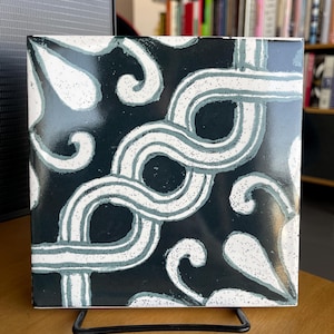 Puede incluir: Un azulejo de cerámica blanco y negro con un patrón repetitivo de espirales estilizados y formas de lágrima. El azulejo está sobre un soporte negro.