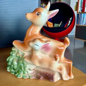 May include: A ceramic figurine of a mother deer and fawn, in shades of tan, pink, and white. The deer are posed together, with the fawn nestled against the mother. The base features green foliage and a brown log.