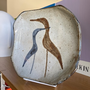 May include: A speckled, off-white ceramic plate with an irregular shape. The plate features a stylized illustration of two birds, one brown and one blue. The birds are depicted with long necks and legs. The plate has a slightly raised edge.