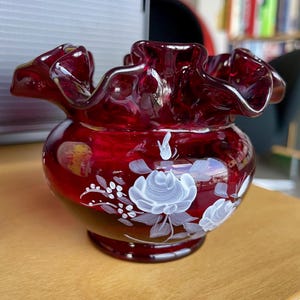 Może przedstawiać: Rubinowoczerwony szklany wazon z falbanką i okrągłym korpusem. Wazon jest ozdobiony białymi wzorami kwiatowymi. Wazon stoi na drewnianej powierzchni.