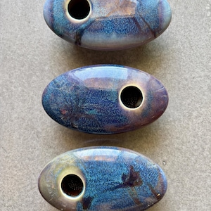 May include: Three oval-shaped ceramic vases with a glossy, iridescent finish in shades of blue, purple, and bronze. Each vase has a single, large circular opening. The vases are arranged vertically.