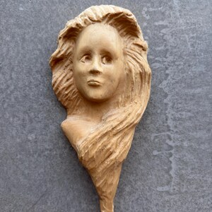 May include: A light brown wooden carving of a human head and shoulders featuring flowing, textured hair that frames the face. The sculpture displays visible chisel marks throughout the surface, creating a detailed representation of facial features.