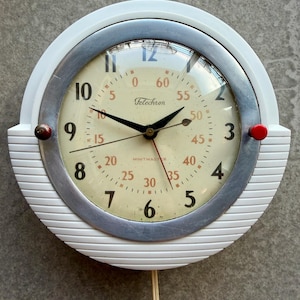 以下が含まれることがあります： ヴィンテージの白とシルバーのテレクロン壁掛け時計。文字盤はクリーム色で、黒い数字と針が付いています。側面に赤いアクセントがあります。時計の文字盤には「Telechron」と「MINITMASTER」の文字があります。