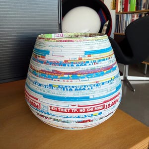 May include: A decorative, handcrafted vase made from layered paper strips. The vase features a cylindrical shape with a wide opening. The paper strips display colorful text and patterns, including the text "TIEN THIET TP. HO CHI MINH 10.000".