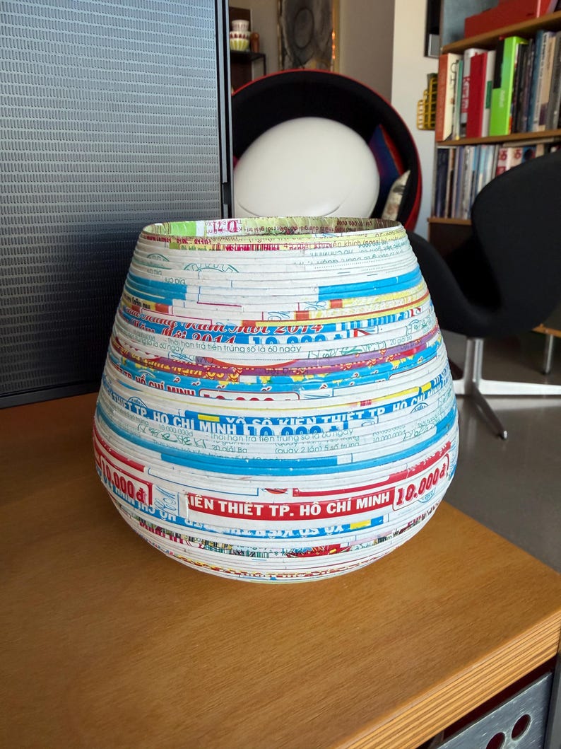May include: A decorative, handcrafted vase made from layered paper strips. The vase features a cylindrical shape with a wide opening. The paper strips display colorful text and patterns, including the text "TIEN THIET TP. HO CHI MINH 10.000".