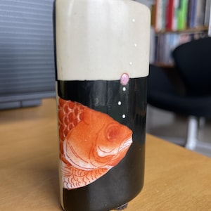 May include: A ceramic vase with a white top and a black base. An orange koi fish design is on the black portion. The vase has a rounded rectangular shape and small white dots and a pink dot on the white portion.