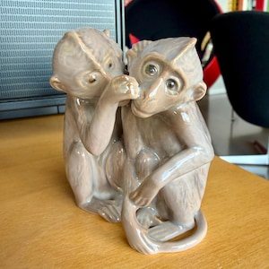 May include: A ceramic figurine depicting two monkeys in a light brown color. One monkey covers its mouth with a hand, while the other looks on. The figurine is placed on a wooden surface.