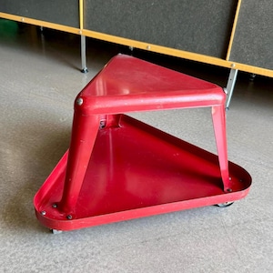 May include: A red, triangular metal cart with a tray base and a three-legged frame. The cart has small wheels for easy movement. The design is minimalist and functional, suitable for various uses.