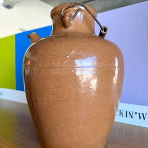 以下が含まれることがあります： ハンドルと金属ワイヤーの留め具が付いた茶色のセラミックジャグ。ジャグは丸みを帯びたボディとテクスチャード加工された表面を持っています。背景には棚とカラフルな壁パネルが含まれています。