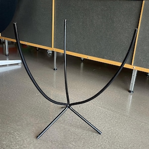 Op de afbeelding: Een zwarte metalen plantenstandaard met drie poten en een gebogen bovenkant. De standaard is ontworpen om een grote plantenpot te houden.