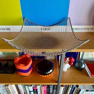 May include: A square, silver-toned wire mesh basket with curved, upward-sweeping edges. The metal grid features a uniform pattern of small squares, creating a shallow, concave basin shape. The structure is supported by a thin, metallic frame.