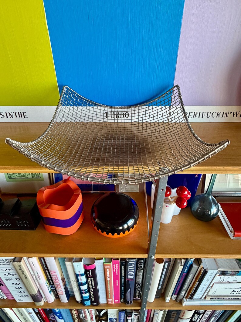 May include: A square, silver-toned wire mesh basket with curved, upward-sweeping edges. The metal grid features a uniform pattern of small squares, creating a shallow, concave basin shape. The structure is supported by a thin, metallic frame.
