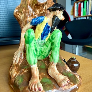 May include: A glazed ceramic figurine depicting a bearded figure with a black hat, blue and yellow shirt, and green pants, sitting against a brown tree stump. A small dark brown round object rests on the ground near the figure's right foot.