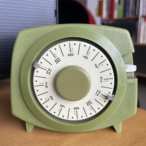 Puede incluir: Un temporizador de plástico verde con una esfera blanca y marcas negras. La esfera está configurada a las 12:00 del mediodía. El temporizador tiene una perilla en el centro y una palanca en el lateral.