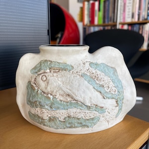 May include: A white ceramic vase with a textured surface and a raised design of a fish in a green and white pattern.