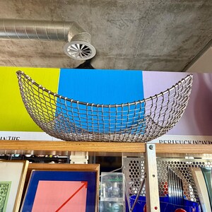 May include: A curved, silver-toned metal wire mesh basket with an open-weave grid pattern. The basket features a wide, shallow, crescent-like shape with a reinforced rim, designed for holding items while maintaining a lightweight, industrial appearance.