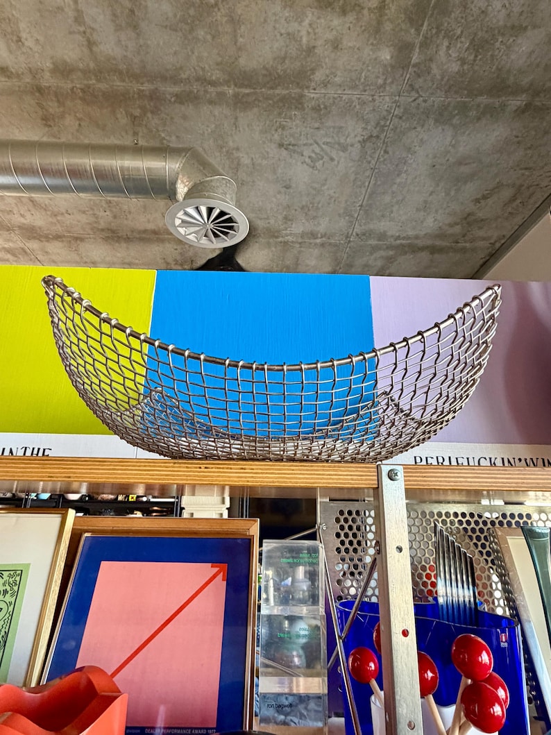 May include: A curved, silver-toned metal wire mesh basket with an open-weave grid pattern. The basket features a wide, shallow, crescent-like shape with a reinforced rim, designed for holding items while maintaining a lightweight, industrial appearance.