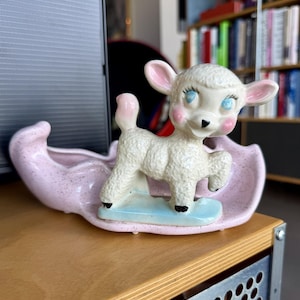 May include: A vintage ceramic lamb figurine with blue eyes, pink cheeks, and a pink tail, standing on a light blue base. The lamb is set in a pink planter with a speckled design. A charming decorative piece.